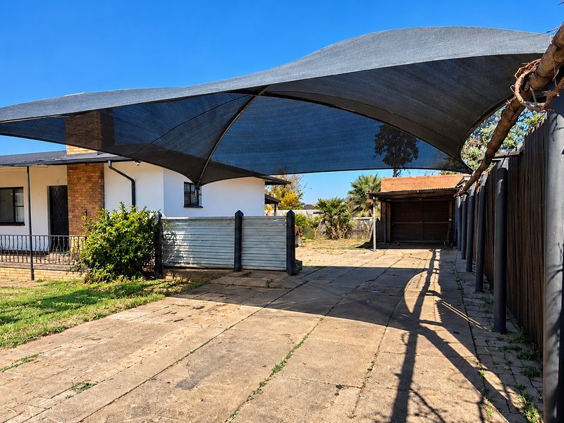 Front exterior view featuring a paved driveway with a large shade sail, brick house facade, and a detached metal garage structure.