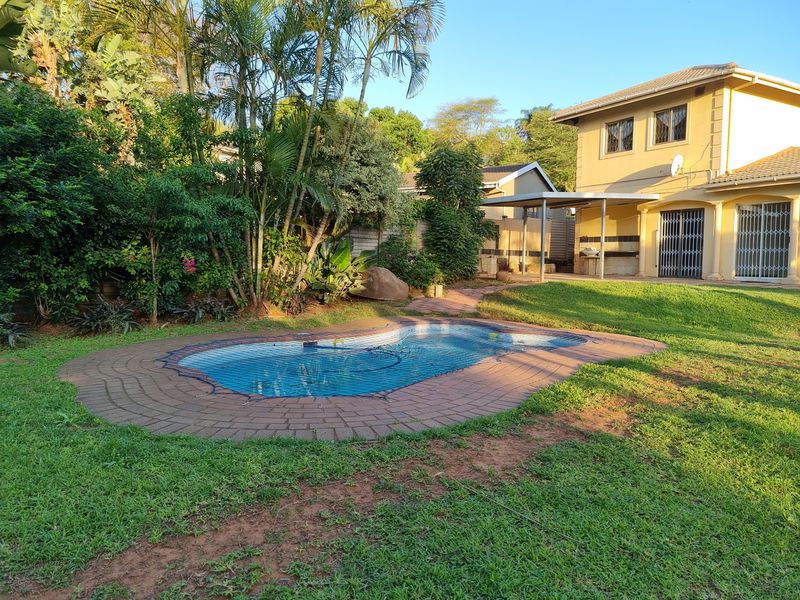A backyard featuring a swimming pool with brick coping, a covered patio area, and a well-established garden with mature trees.