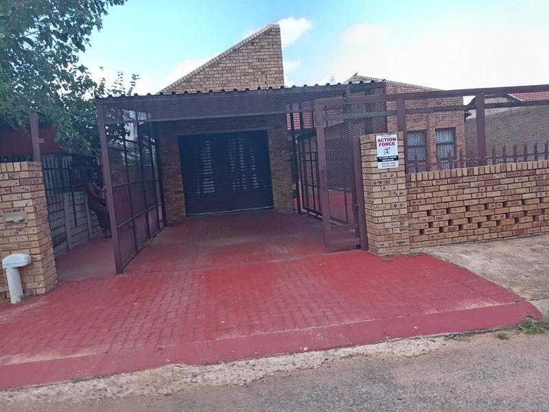 Front exterior view of a face brick house featuring a paved driveway, metal carport, and secure perimeter walling with gated access.