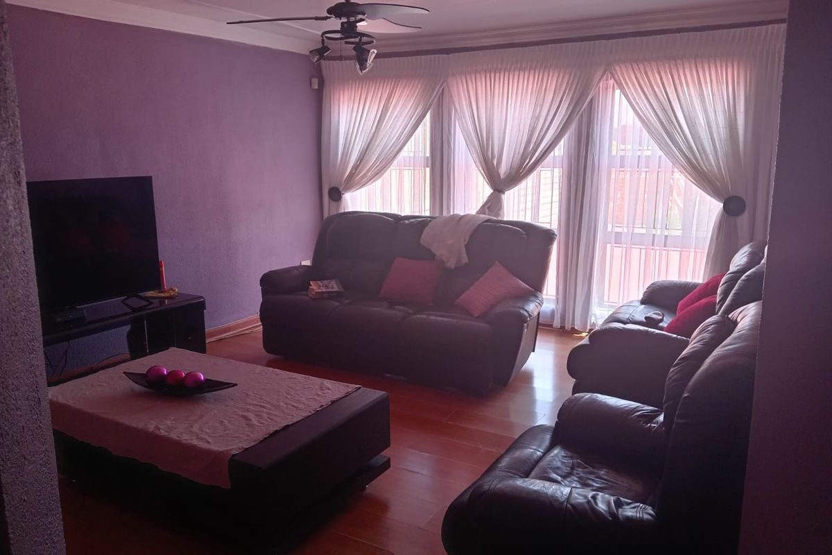 A comfortable living room featuring laminate flooring, a ceiling fan, and large windows with draped curtains.
