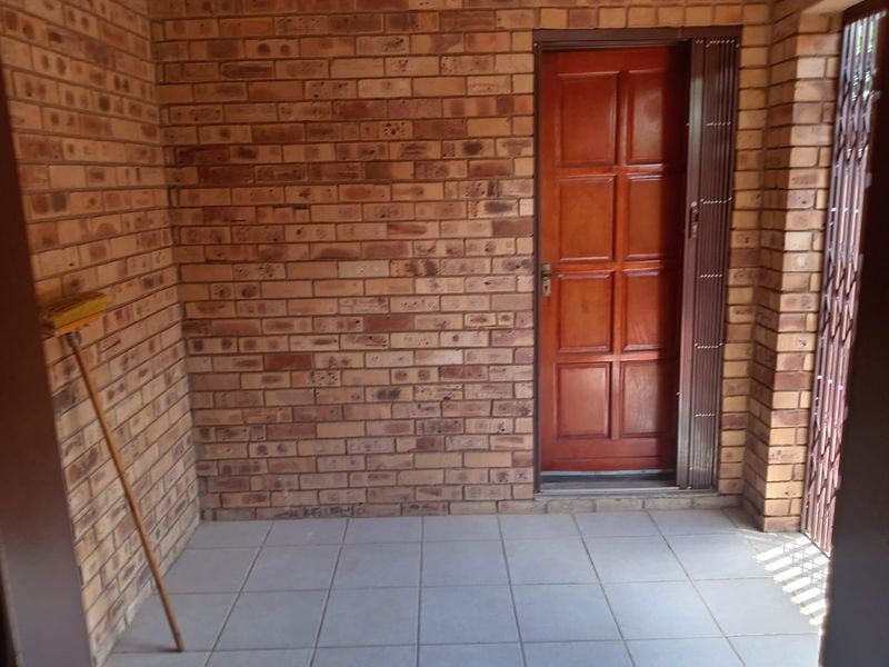 A covered patio area featuring face brick walls, tiled flooring, and a secure timber entrance door with a metal security gate.