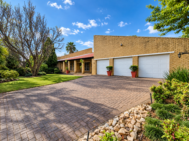 A well-maintained brick house featuring a spacious triple garage, a neatly paved driveway, and an established garden with mature trees.