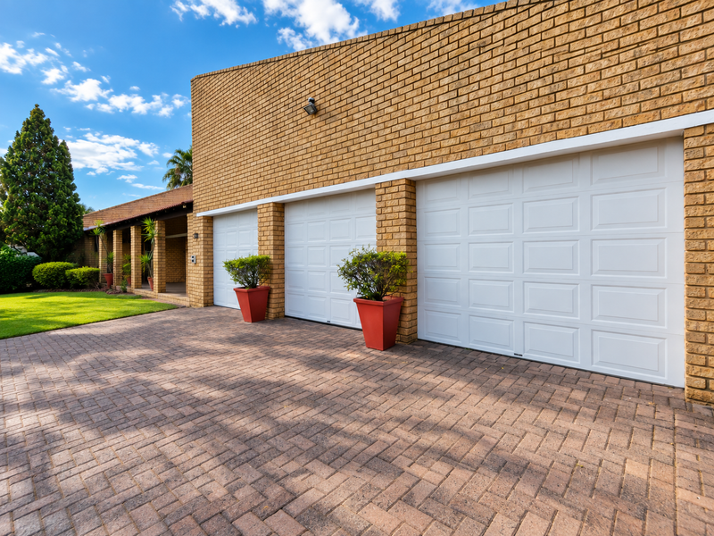 A well-maintained front exterior featuring a triple garage with white sectional doors, a paved driveway, and a covered patio area.