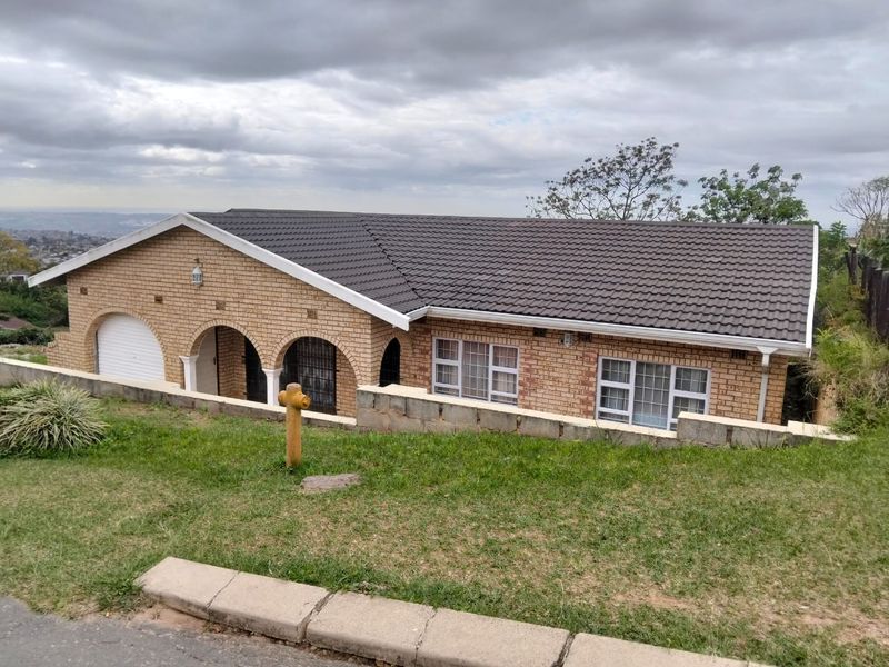 Front elevation of a face-brick house featuring a tiled roof, arched entrance, and integrated garage, set on a sloped plot with a retaining wall.