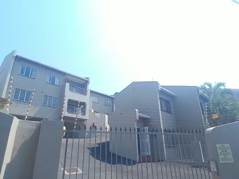 Front exterior view of a townhouse complex featuring a gated entrance, paved driveway, and multi-story residential units with balconies.