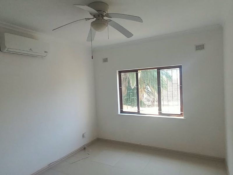 A bright, empty bedroom featuring tiled flooring, a ceiling fan, and a wall-mounted split-system air conditioner.