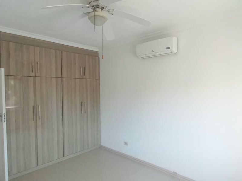 A bright, empty bedroom featuring built-in wooden cupboards, a ceiling fan, and a wall-mounted air conditioning unit, with an adjoining bathroom visible.