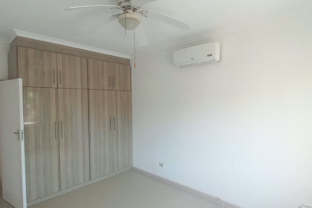 A bright, empty bedroom featuring built-in wooden cupboards, a ceiling fan, and a wall-mounted air conditioning unit, with an adjoining bathroom visible.