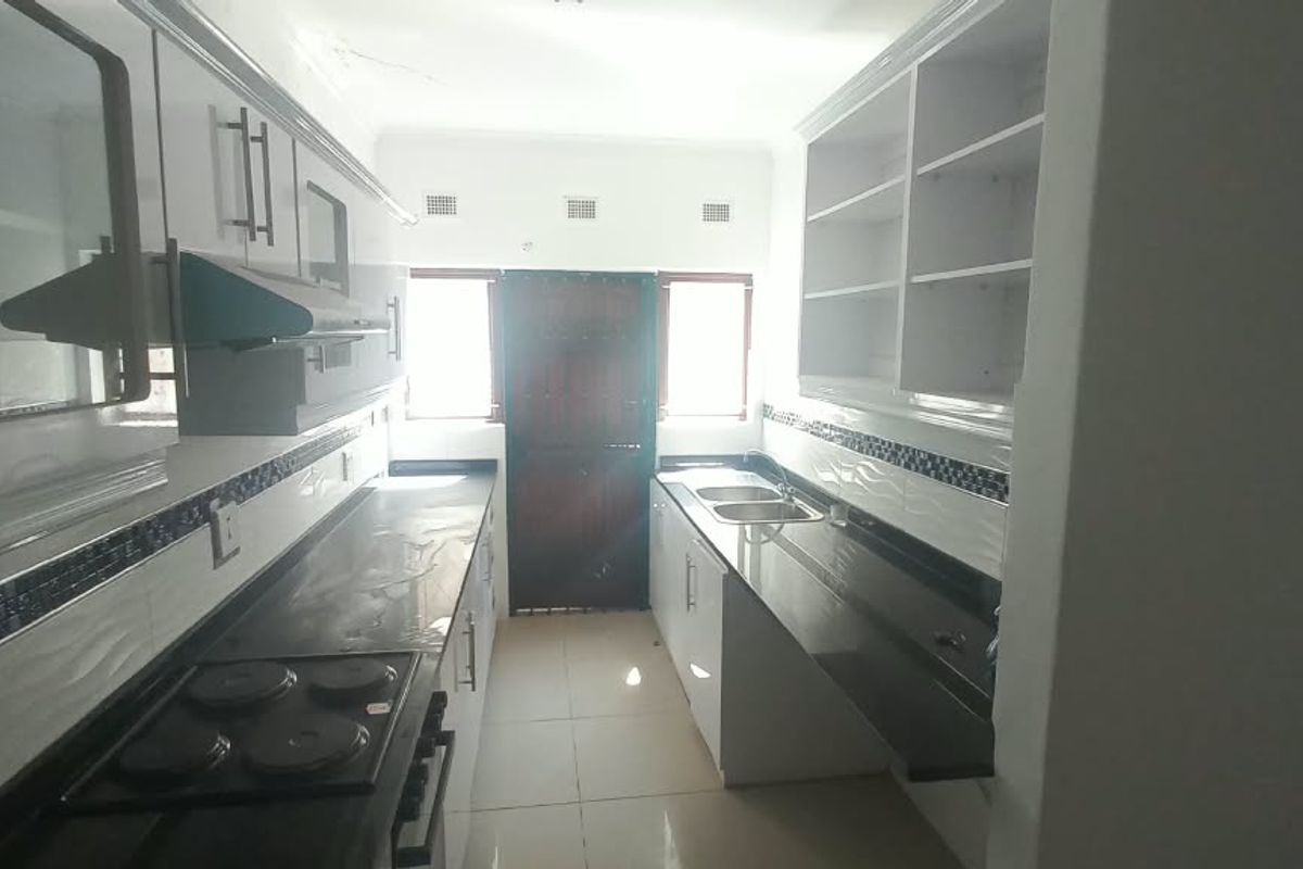 A functional galley-style kitchen featuring built-in cabinetry, dark countertops, an electric hob, and a double sink with tiled backsplash.