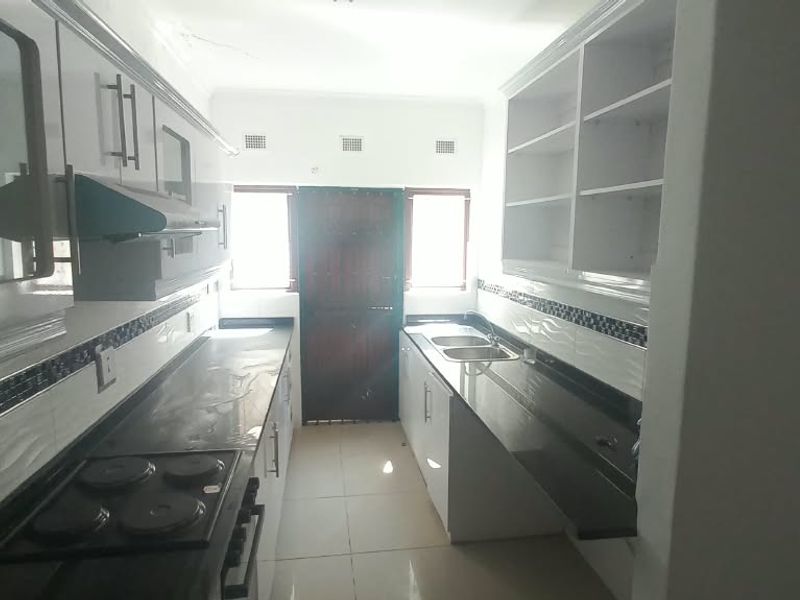 A functional galley-style kitchen featuring built-in cabinetry, dark countertops, an electric hob, and a double sink with tiled backsplash.