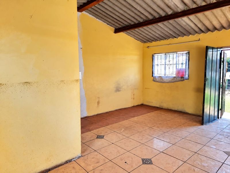 An empty living area featuring tiled flooring, exposed roof beams, and security bars on the window, requiring cosmetic updates and wall repairs.