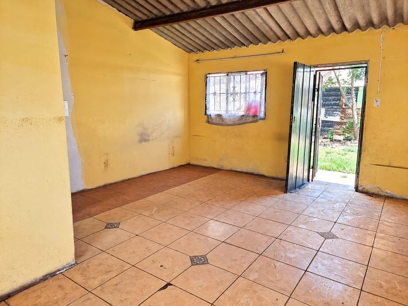 An empty living area featuring tiled floors, exposed roof structure, and direct access to the garden through a hinged door.