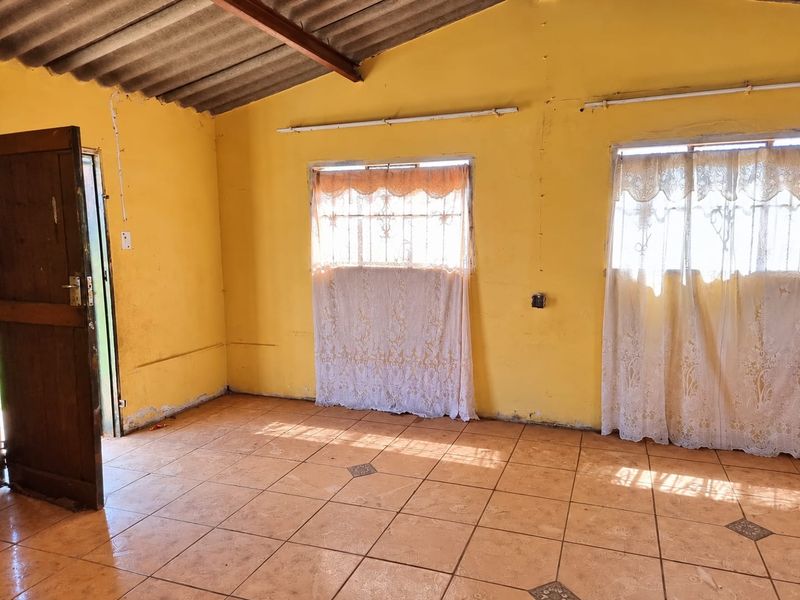 An empty living room featuring tiled floors, exposed rafters, and security-barred windows, requiring cosmetic updates and wall repairs to improve its overall condition.