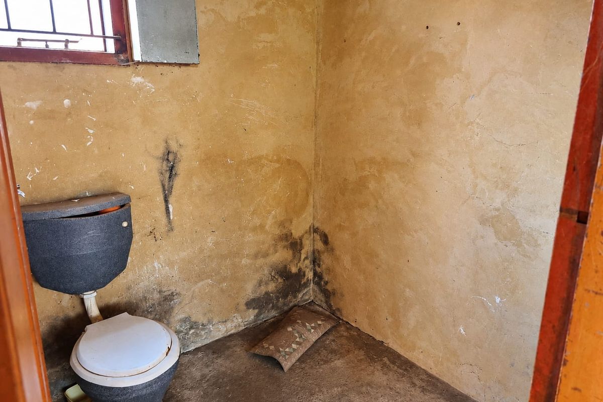 A small, basic bathroom requiring significant renovation, featuring a toilet and security bars on the window.