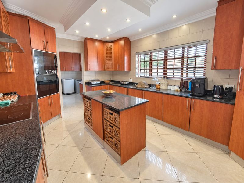 Spacious kitchen featuring warm wood cabinetry, granite countertops, a central island, and integrated appliances, with a separate scullery area visible in the background.