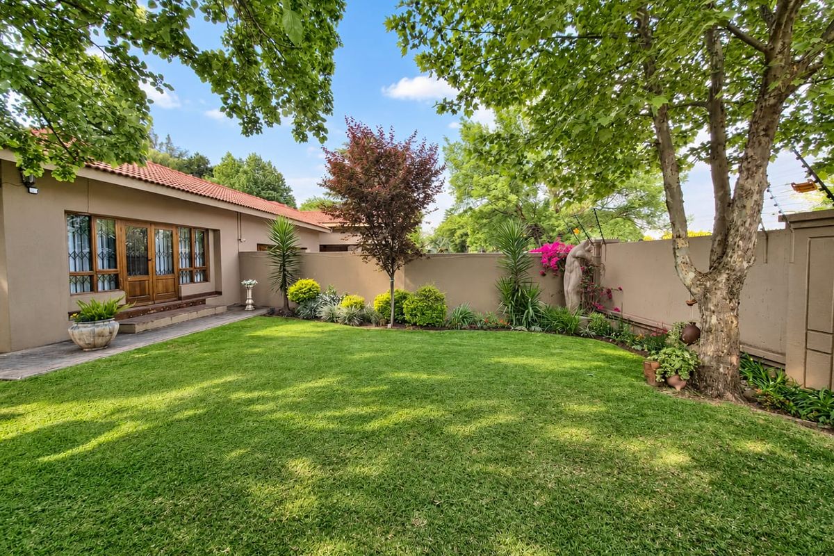 A well-maintained rear garden featuring a lush lawn, mature trees, perimeter wall with electric fencing, and direct access to the home via timber-framed doors.