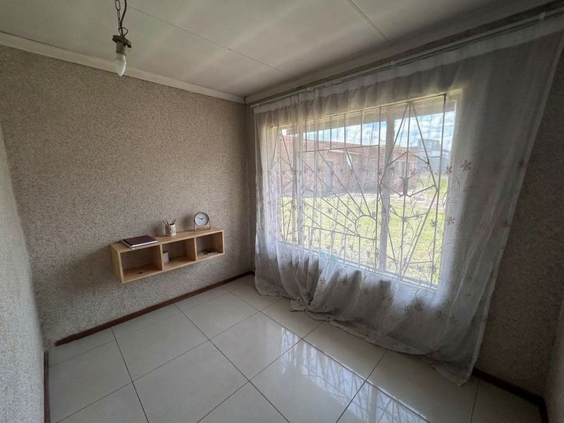 A compact bedroom featuring tiled flooring, a large window with security bars, and a built-in wall shelf, offering a blank canvas for renovation.