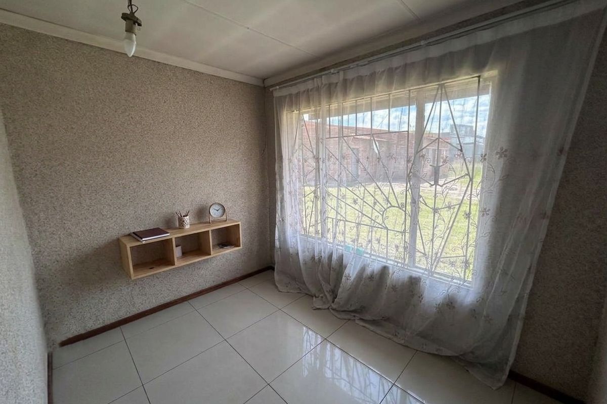 A compact bedroom featuring tiled flooring, a large window with security bars, and a built-in wall shelf, offering a blank canvas for renovation.