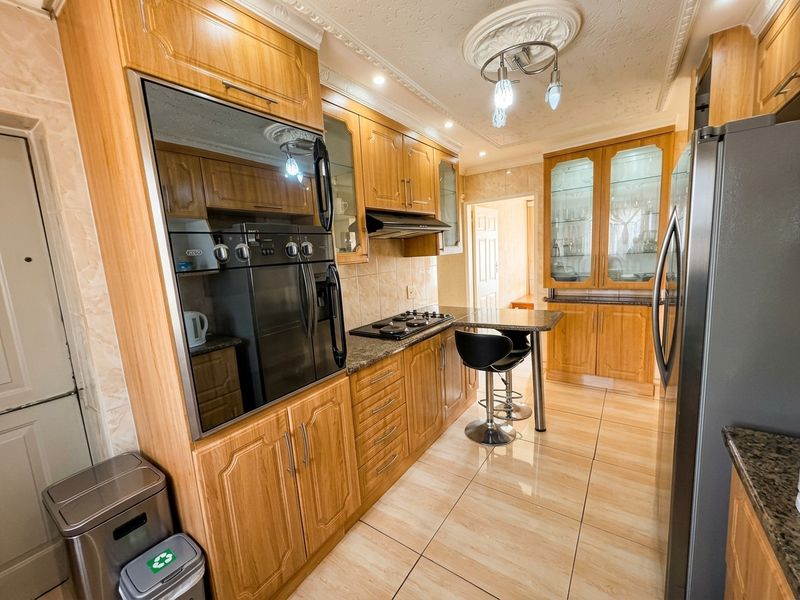 A well-maintained kitchen featuring warm-toned wooden cabinetry, granite countertops, built-in oven, gas hob, and elegant glass-fronted display units with tiled flooring.