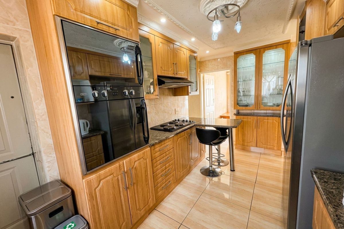 A well-maintained kitchen featuring warm-toned wooden cabinetry, granite countertops, built-in oven, gas hob, and elegant glass-fronted display units with tiled flooring.