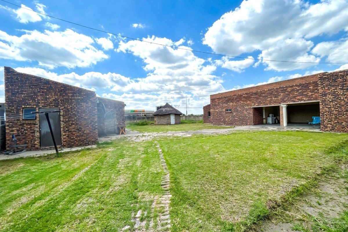 Spacious rear yard featuring exposed brick structures, a traditional thatched rondavel, and a covered patio area set within a large lawn.