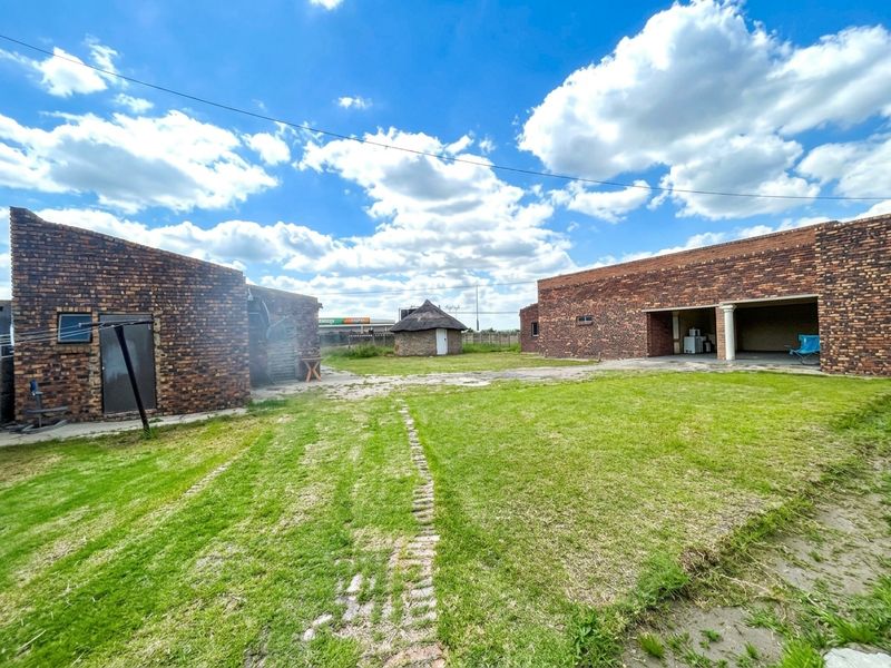 Spacious rear yard featuring exposed brick structures, a traditional thatched rondavel, and a covered patio area set within a large lawn.