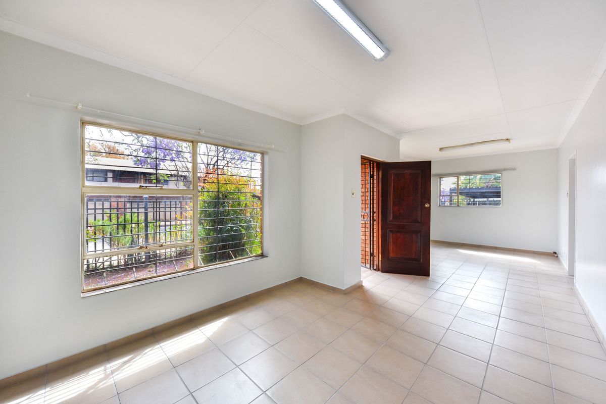 A bright, vacant living area featuring neutral tiled flooring, large windows with security bars, and direct access to an adjacent room.