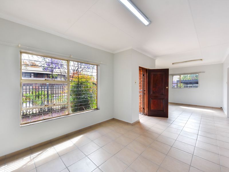 A bright, vacant living area featuring neutral tiled flooring, large windows with security bars, and direct access to an adjacent room.