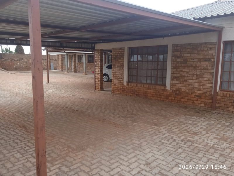 A spacious paved outdoor area featuring a large corrugated metal carport structure attached to an exposed brick building with steel-framed windows.