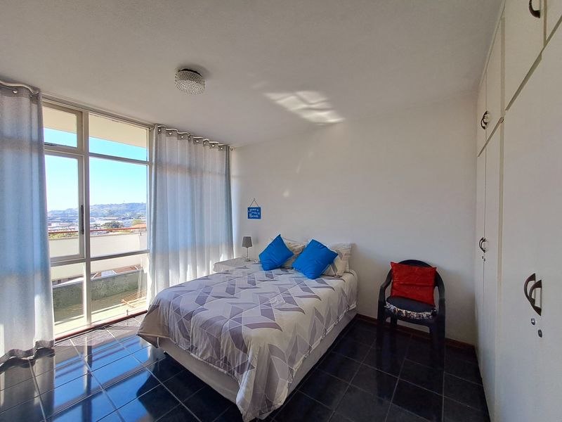 A bright bedroom featuring large floor-to-ceiling windows with natural light, dark tiled flooring, and built-in storage cupboards.