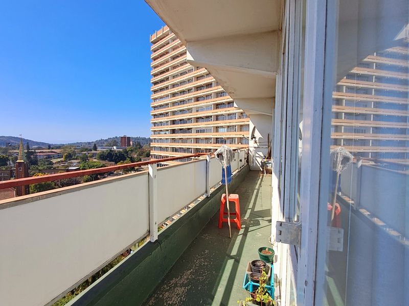 A spacious apartment balcony featuring a metal railing and sliding glass doors, offering an elevated urban view with potential for cosmetic refurbishment.