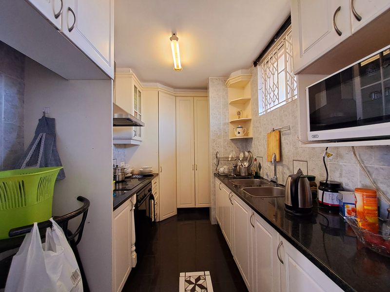 A functional galley-style kitchen featuring white cabinetry, dark countertops, a double basin sink, and built-in appliances including a hob and microwave.