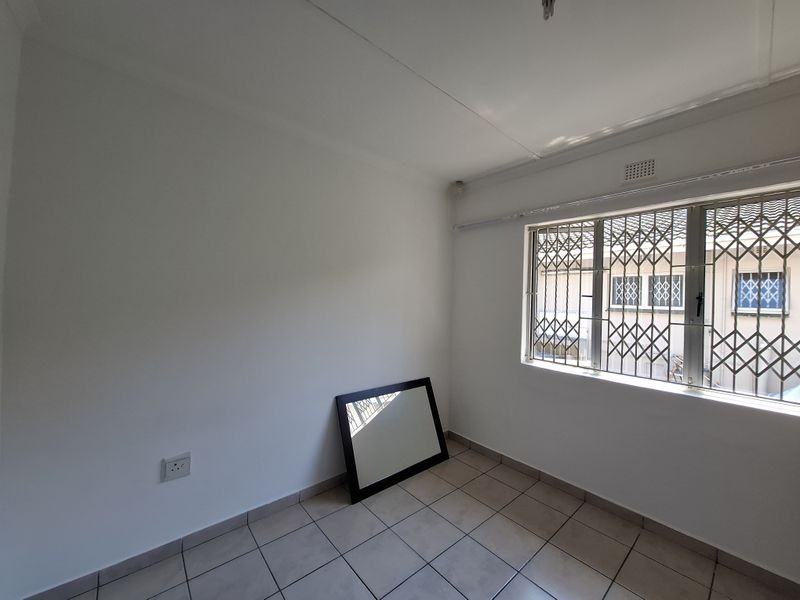 A bright, empty bedroom featuring neutral tiled flooring, white walls, and a window fitted with a security gate.