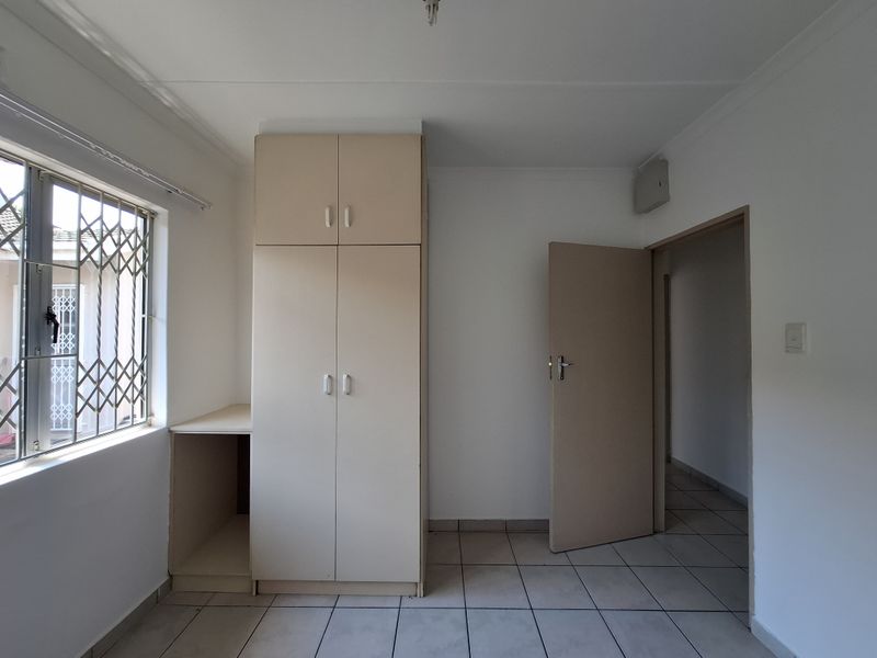 A simple, empty bedroom featuring tiled flooring, built-in cupboards, and a window fitted with security burglar bars.