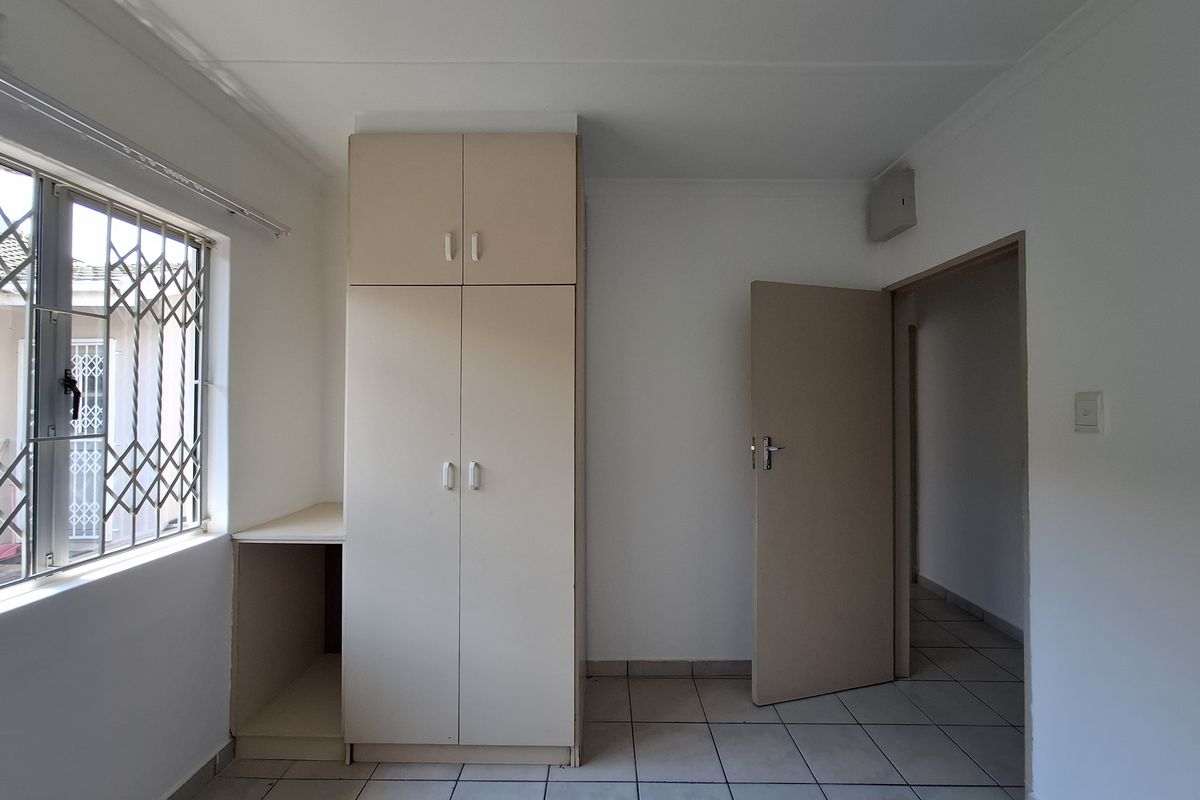 A simple, empty bedroom featuring tiled flooring, built-in cupboards, and a window fitted with security burglar bars.