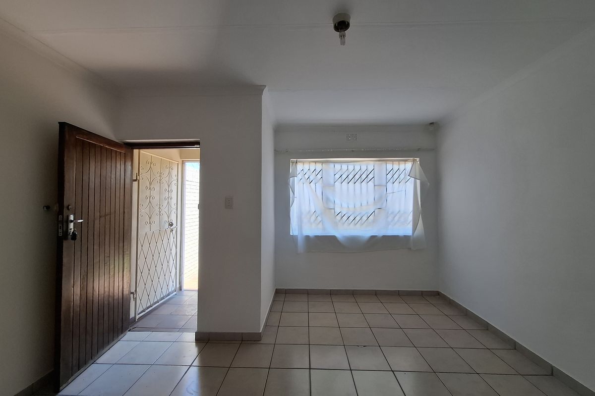 An empty living room featuring tiled flooring, a timber entrance door with a security gate, and a window fitted with security bars.