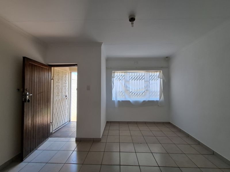 An empty living room featuring tiled flooring, a timber entrance door with a security gate, and a window fitted with security bars.
