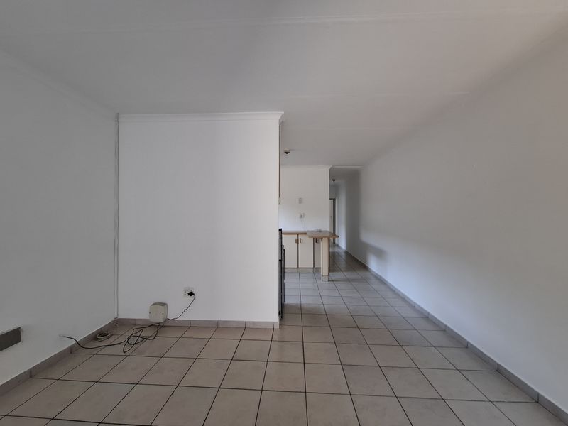 An open-plan living and kitchen area featuring neutral tiled flooring, built-in kitchen cabinetry, and a breakfast counter in a clean, vacant space.