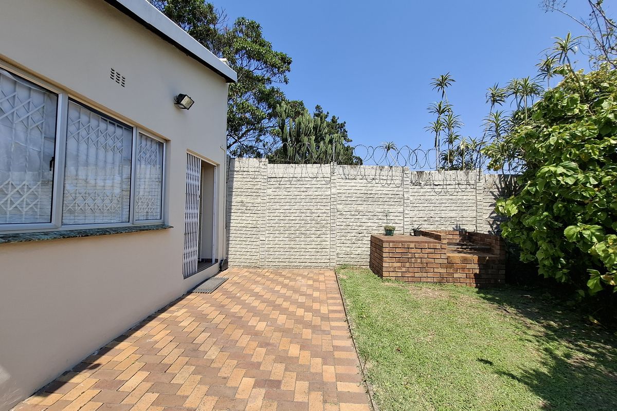 Rear exterior view of the cottage featuring a paved patio area, a built-in brick braai, and a secure perimeter wall with razor wire.