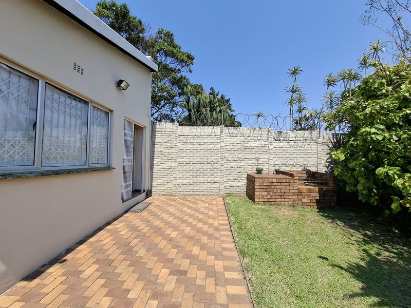 Rear exterior view of the cottage featuring a paved patio area, a built-in brick braai, and a secure perimeter wall with razor wire.