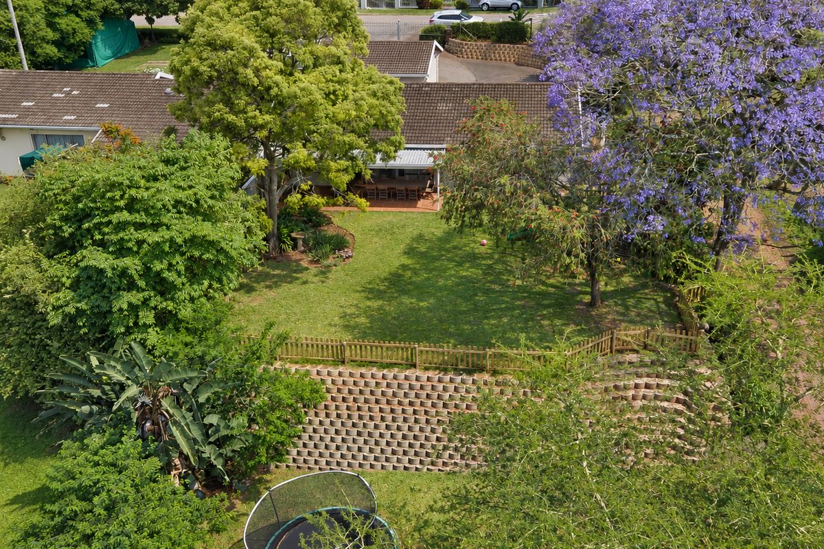 Aerial view of the property featuring a spacious rear garden, established trees, a retaining wall, and a covered patio area.
