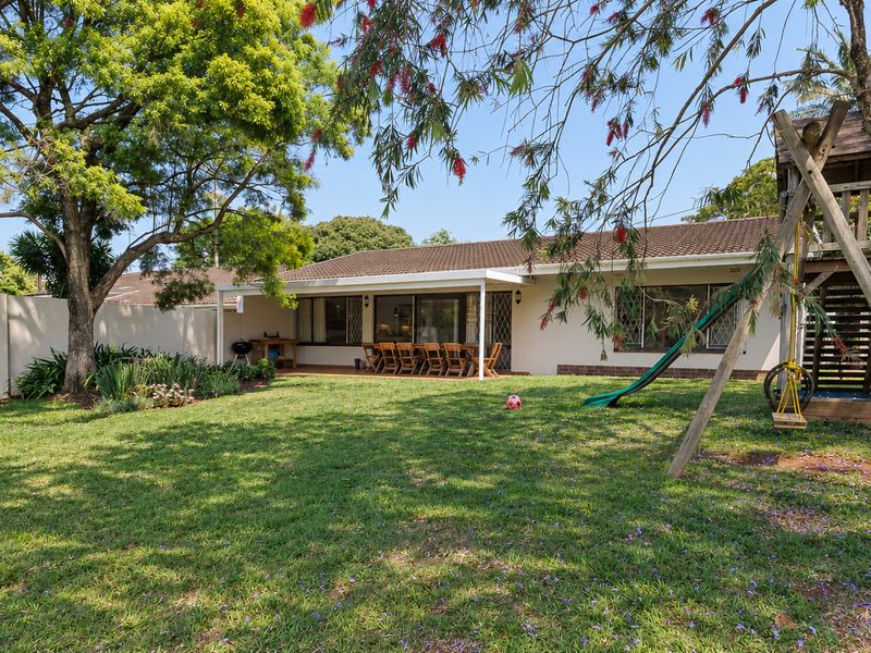 A spacious rear garden featuring a well-maintained lawn, mature trees, and a covered patio area with sliding door access to the main residence.