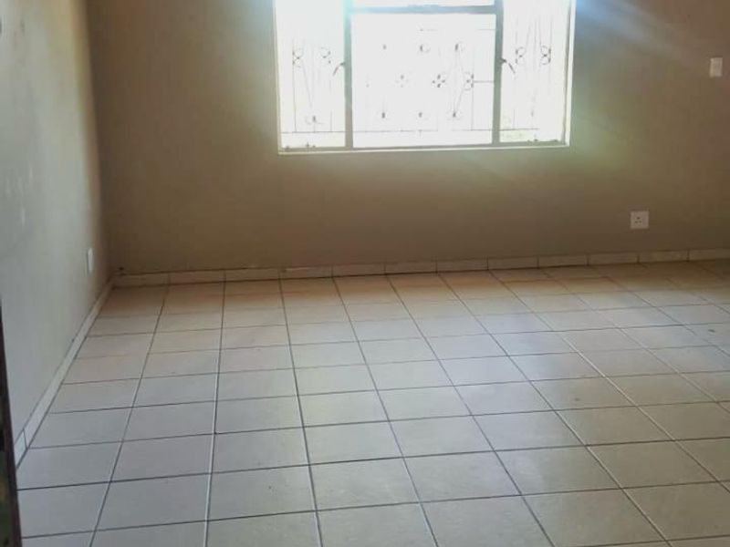 An empty living room featuring ceramic floor tiles, exposed timber ceiling beams, and a window fitted with security burglar bars.