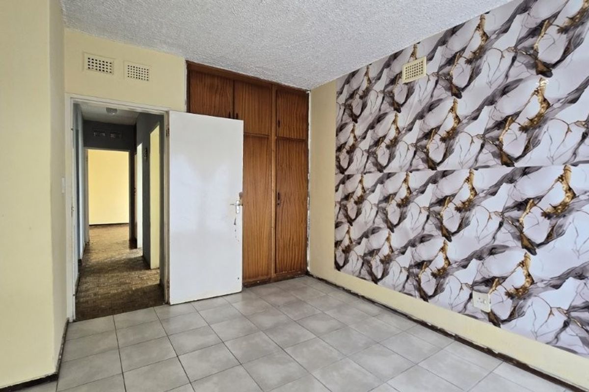 An empty bedroom featuring built-in wooden cupboards, tiled flooring, and a decorative feature wall, offering a blank canvas for renovation.