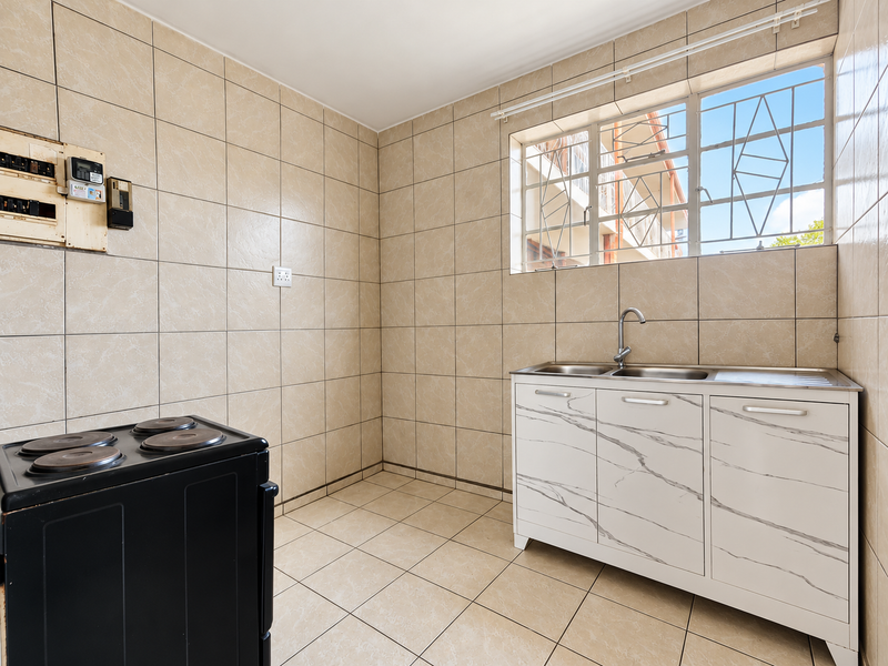 A functional kitchen area featuring fully tiled walls and floors, a stainless steel sink unit with a mixer tap, and a window with security bars.