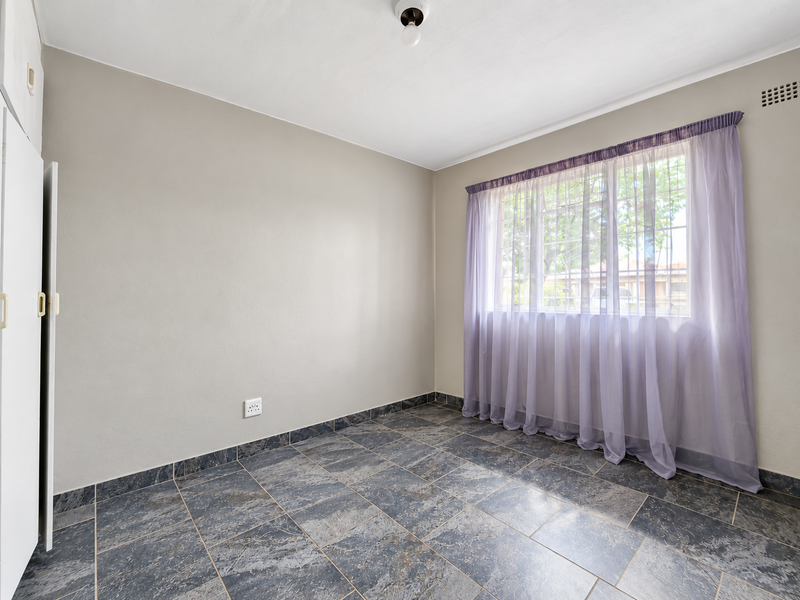 A bright, empty bedroom featuring durable tiled flooring, built-in cupboard storage, and a large window allowing for natural light.