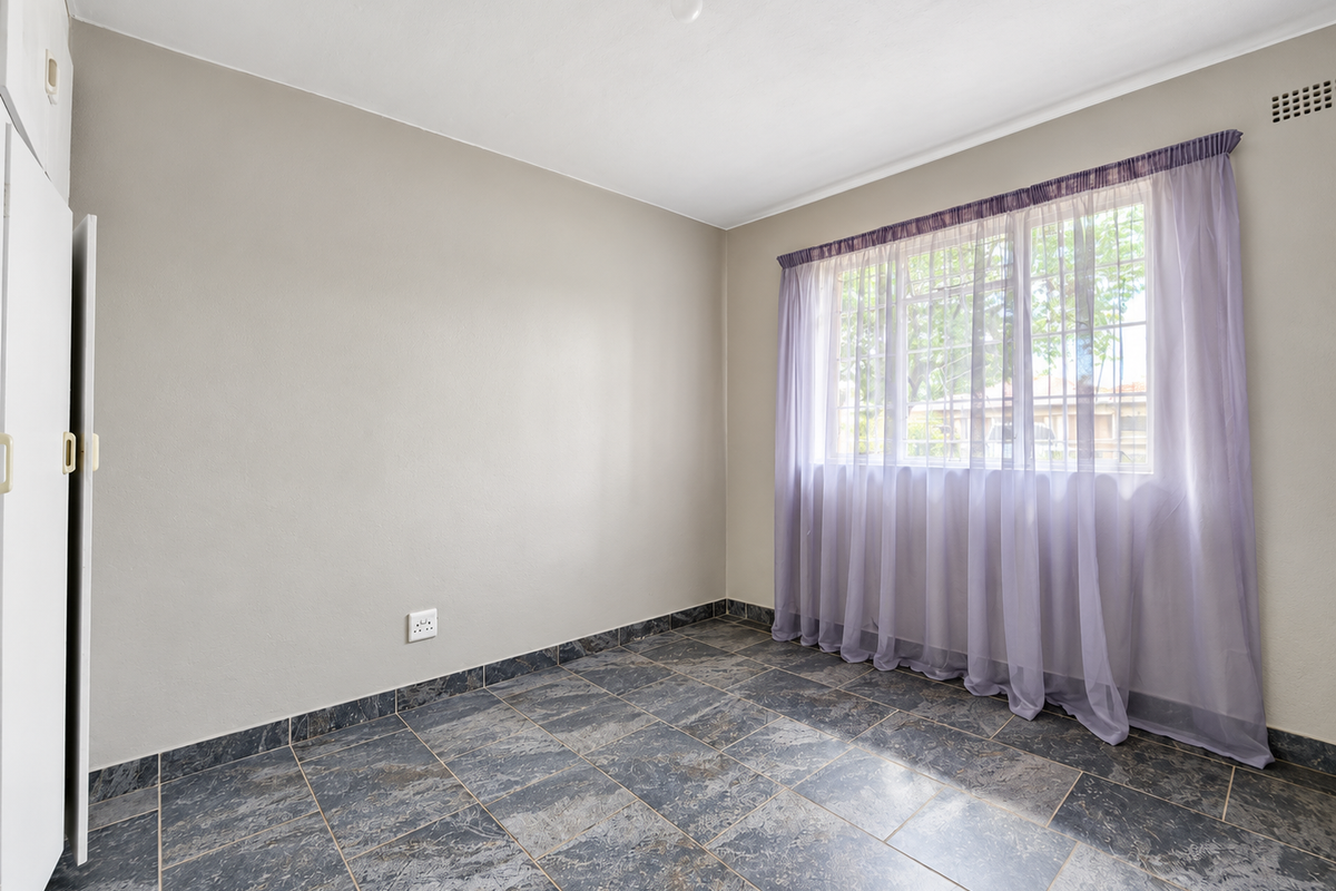 A bright, empty bedroom featuring durable tiled flooring, built-in cupboard storage, and a large window allowing for natural light.