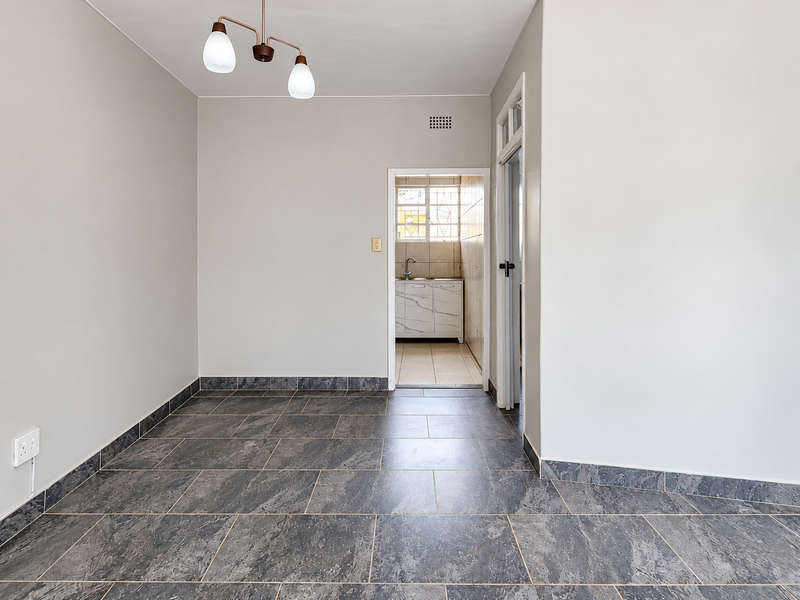 A bright, empty living area featuring modern dark grey floor tiles and neutral walls, with a view into an adjacent scullery area.