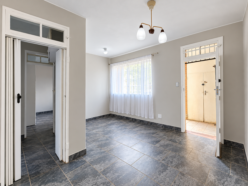 A bright, empty living area featuring neutral walls, dark tiled flooring, and direct access to both an internal hallway and an external entrance.