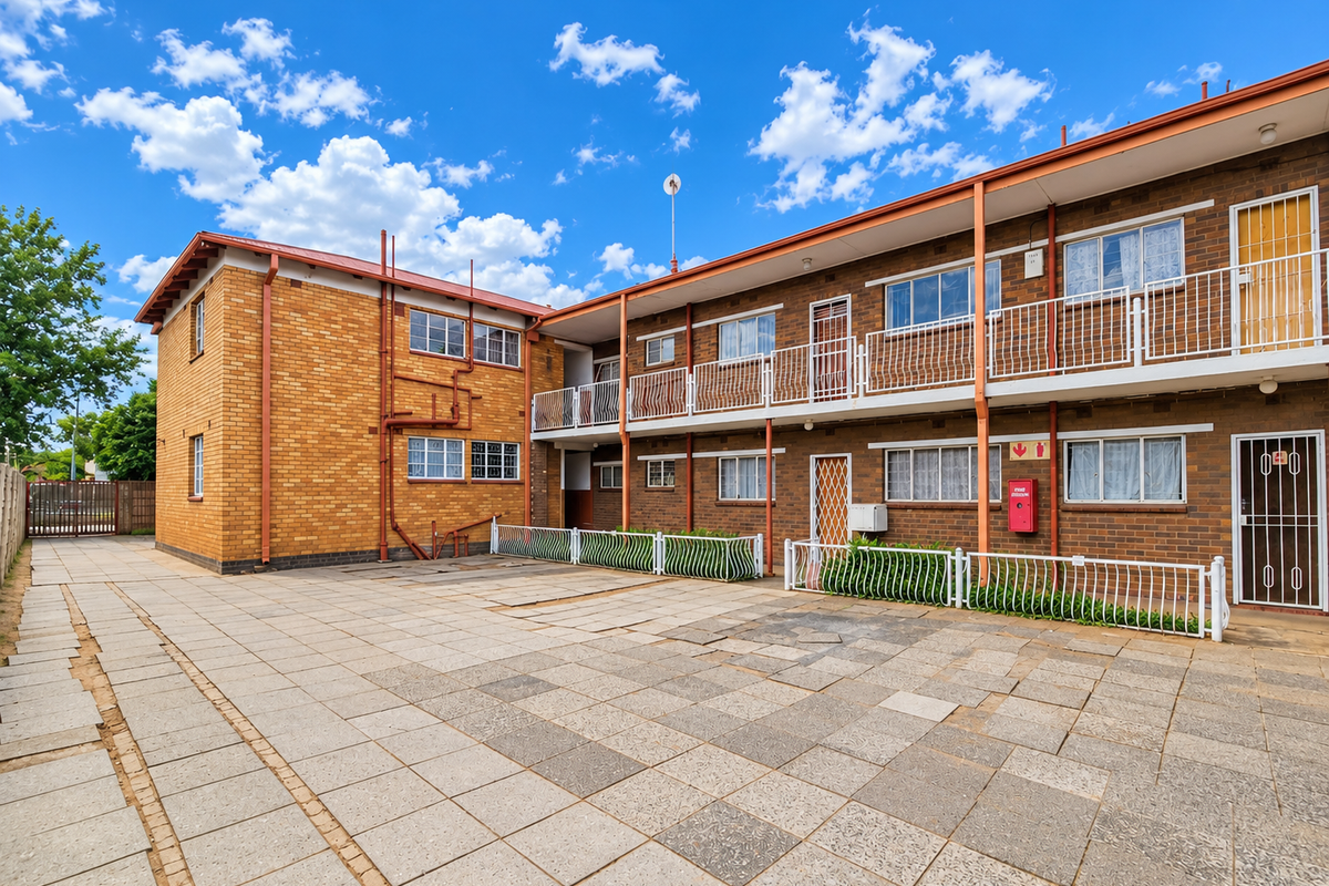 Rear view of a brick apartment building featuring a paved courtyard, metal balcony railings, and security gates on the ground floor units.